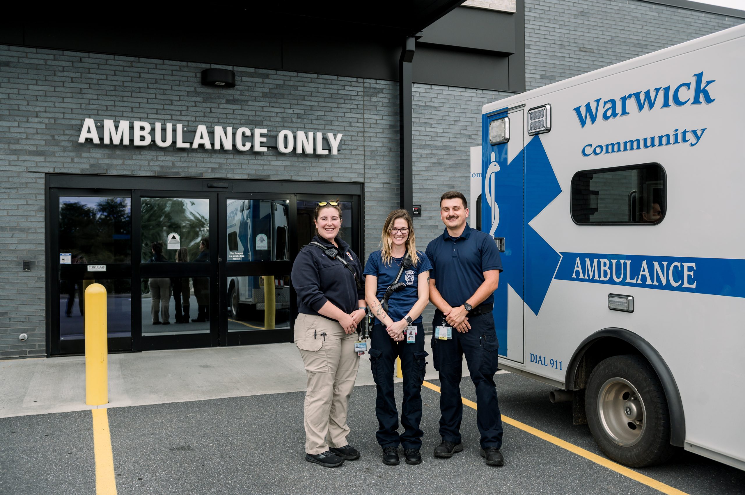 Three Warwick EMS crew members smiling beside a Warwick Community Ambulance at a hospital ambulance entrance.